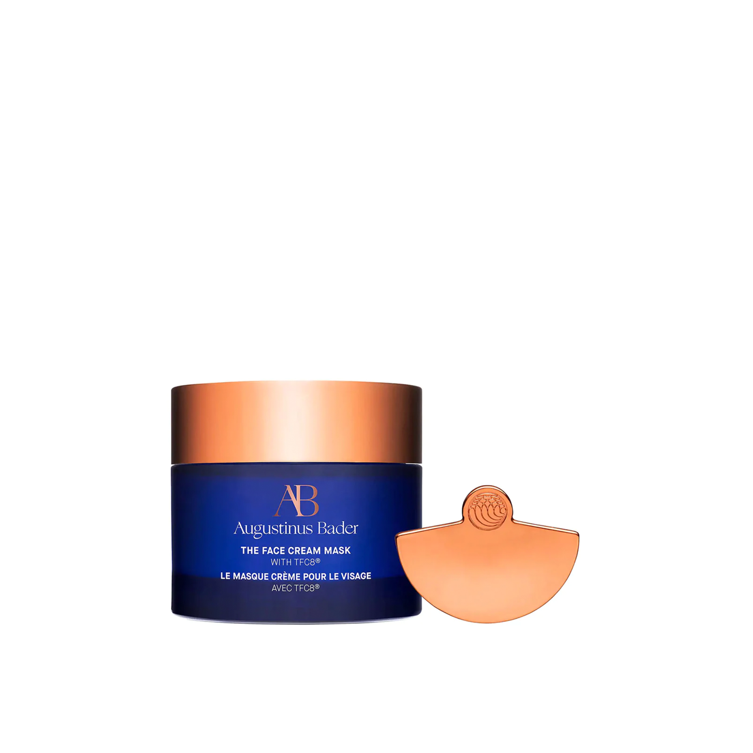 A jar of Augustinus Bader The Face Cream Mask with a copper-colored lid and a provided applicator tool.