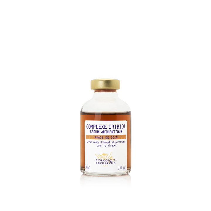 A clear glass vial with a gold cap labeled 'COMPLEXE IRIBIOL' containing a beige-colored liquid, indicative of a skincare serum.